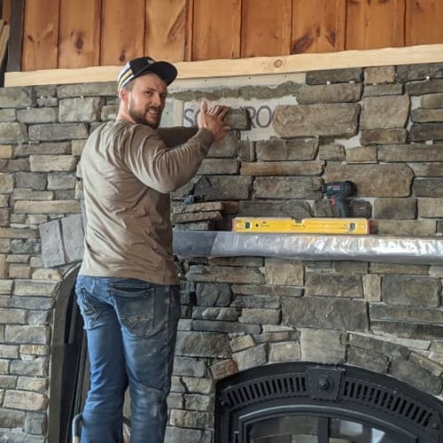 Stone Haven Developments A man in a cap installs stone veneer on a fireplace wall for Stone Haven Developments. Tools and a level rest on the mantel. Ontario