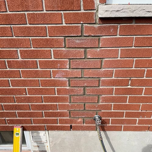 Stone Haven Developments A section of red brick wall by Stone Haven Developments features replacement bricks in the center that don’t match the surrounding ones; a yellow level and a water spigot are visible below. Ontario