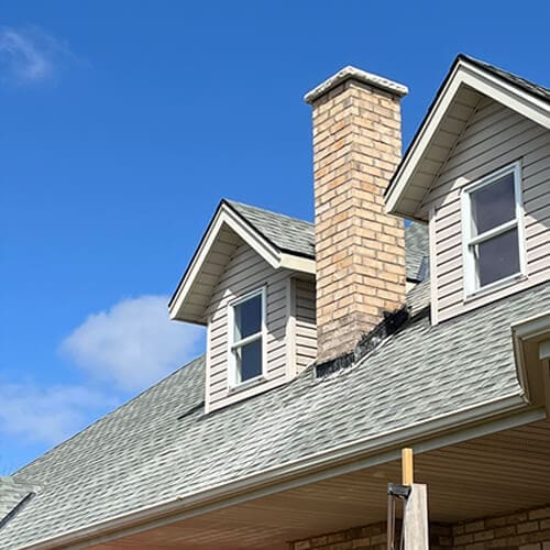 Stone Haven Developments A house roof by Stone Haven Developments features two dormer windows, a brick chimney, and gray shingles under a clear blue sky. Ontario