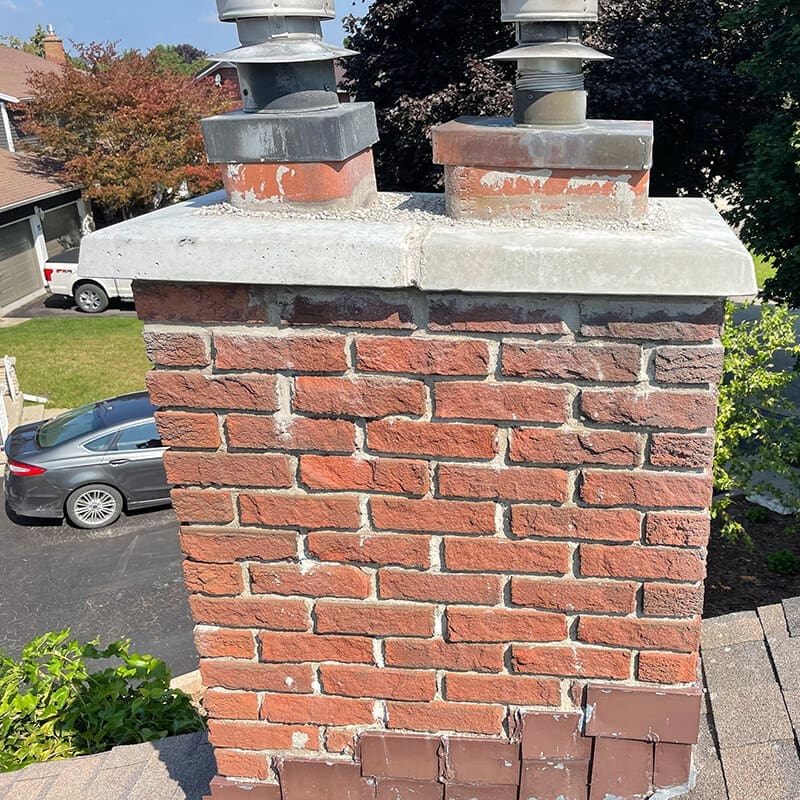 Stone Haven Developments Close-up view of a brick chimney with two metal flue pipes and a concrete cap by Stone Haven Developments, set on a residential rooftop overlooking a driveway and parked car in the background. Ontario