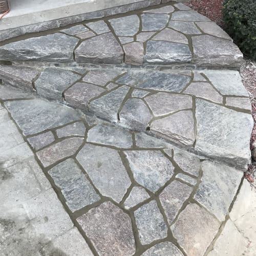 Stone Haven Developments Three outdoor stone steps crafted from irregularly shaped flat stones with visible mortar joints, leading up to a concrete landing—an inviting entrance designed by Stone Haven Developments. Ontario