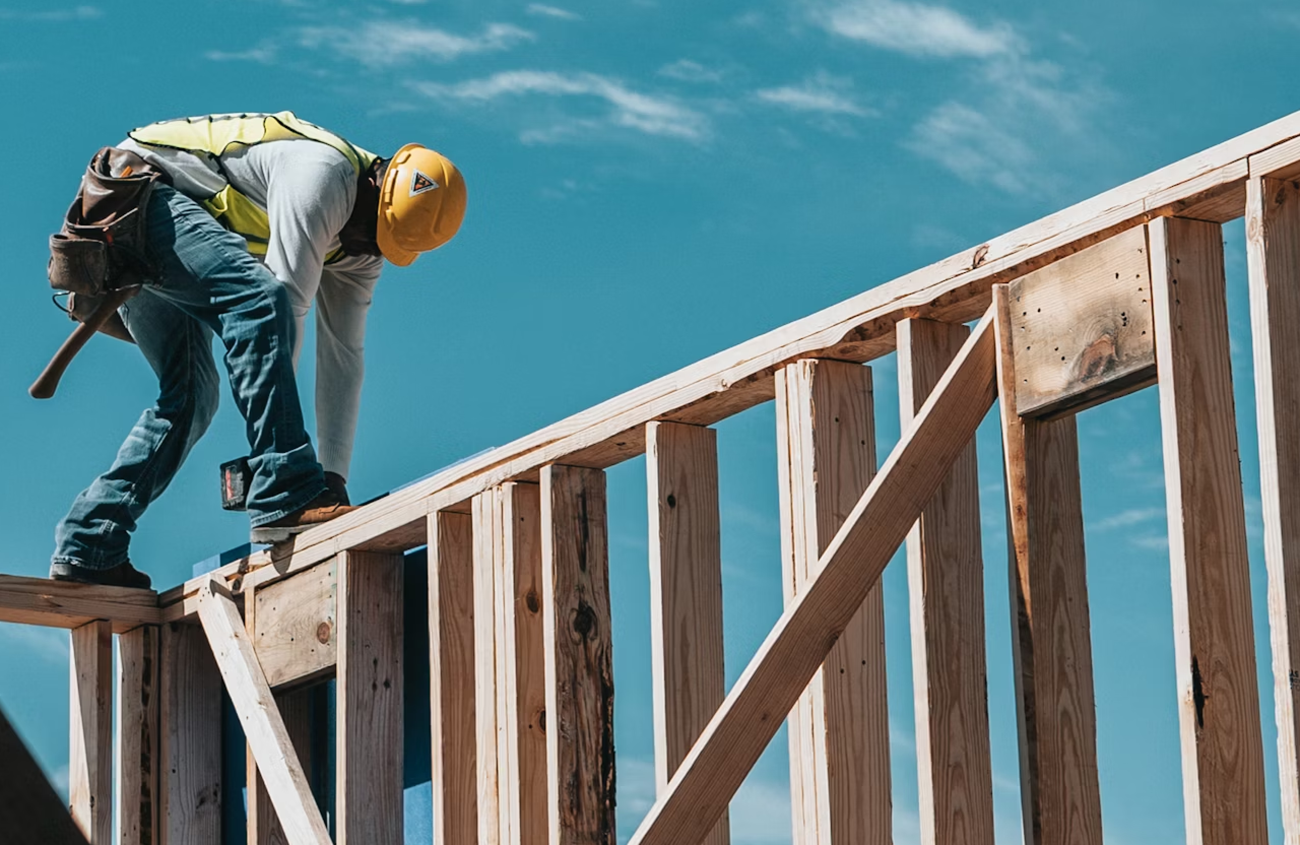 Stone Haven Developments A construction worker in a yellow hard hat and safety vest is building a wooden structure framework under a clear blue sky. Ontario