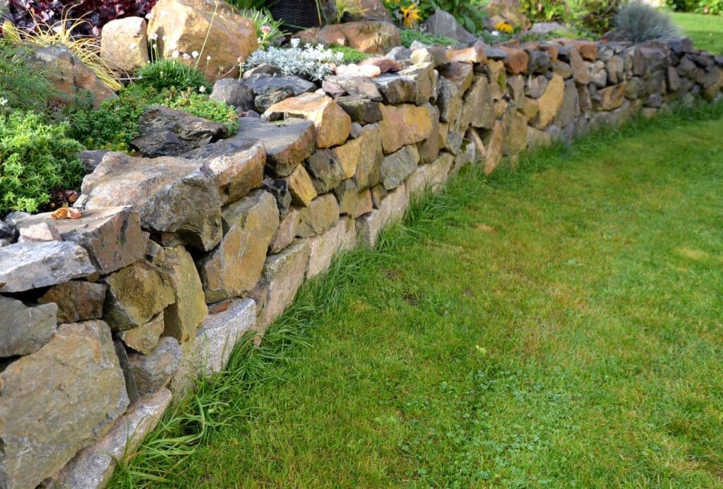 Stone Haven Developments A low stone retaining wall separates a neatly trimmed grassy lawn from a raised garden bed with various green plants. Ontario