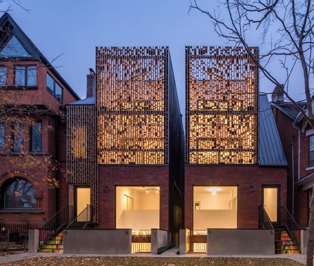 Stone Haven Developments Two modern townhouses with brick facades and decorative metal screens stand side by side between traditional-style homes at dusk. Ontario