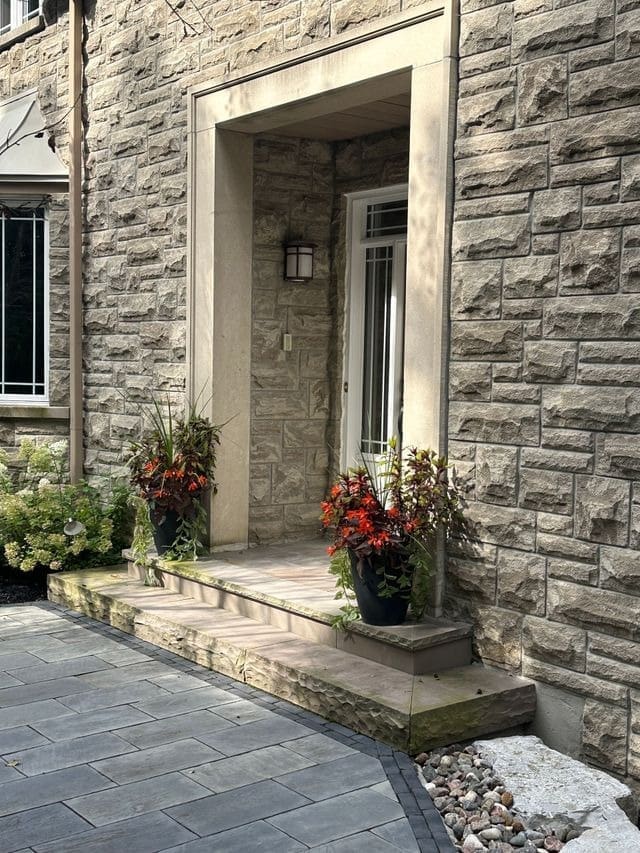 Stone Haven Developments A stone house entrance with a small porch, two potted plants with red flowers on each side of the door, and a glass door with a wall-mounted light above. Ontario