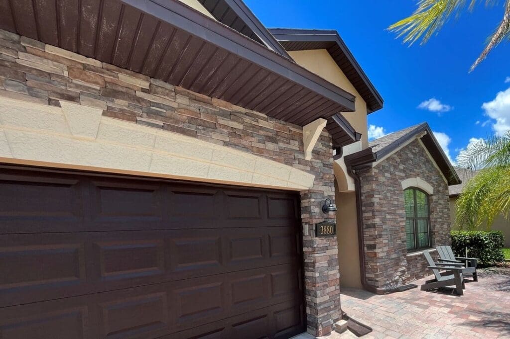 Stone Haven Developments A modern house with a brown brick exterior, a large dark garage door, a bench near the window, and palm trees under a clear blue sky. Ontario