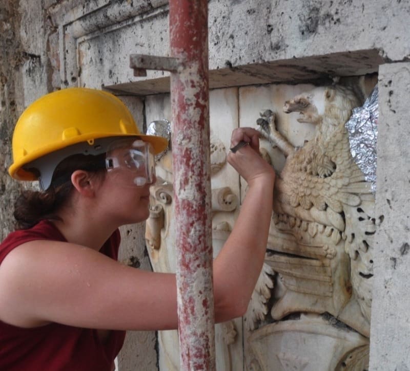 Stone Haven Developments Person wearing a yellow hard hat and safety goggles uses a tool to restore a stone relief sculpture on an exterior wall. Ontario