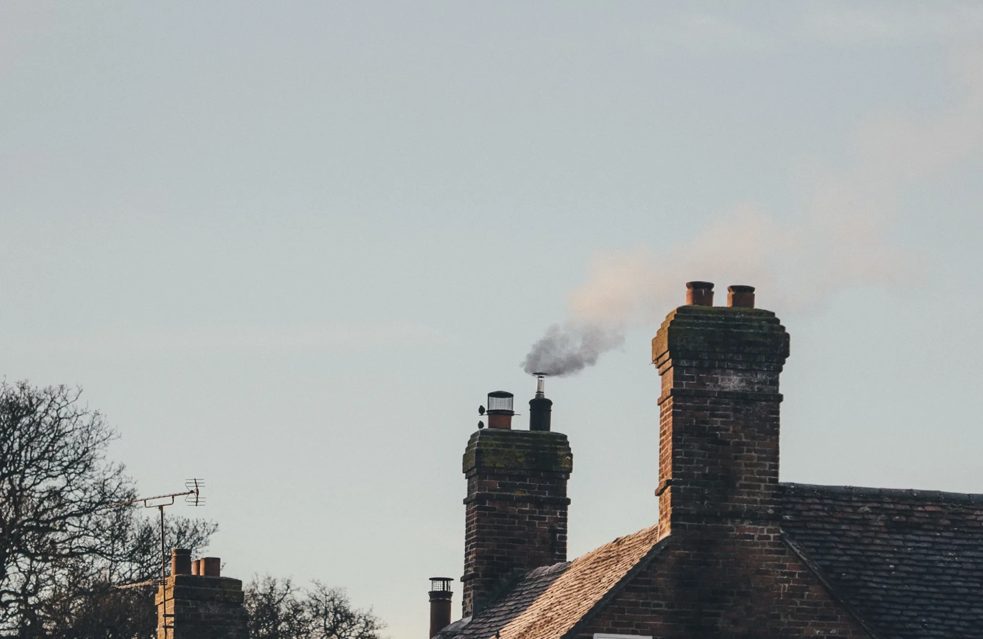 Stone Haven Developments Brick house roof with three chimneys, one actively releasing smoke. Sky is clear, with some trees and a TV antenna visible in the background. Ontario