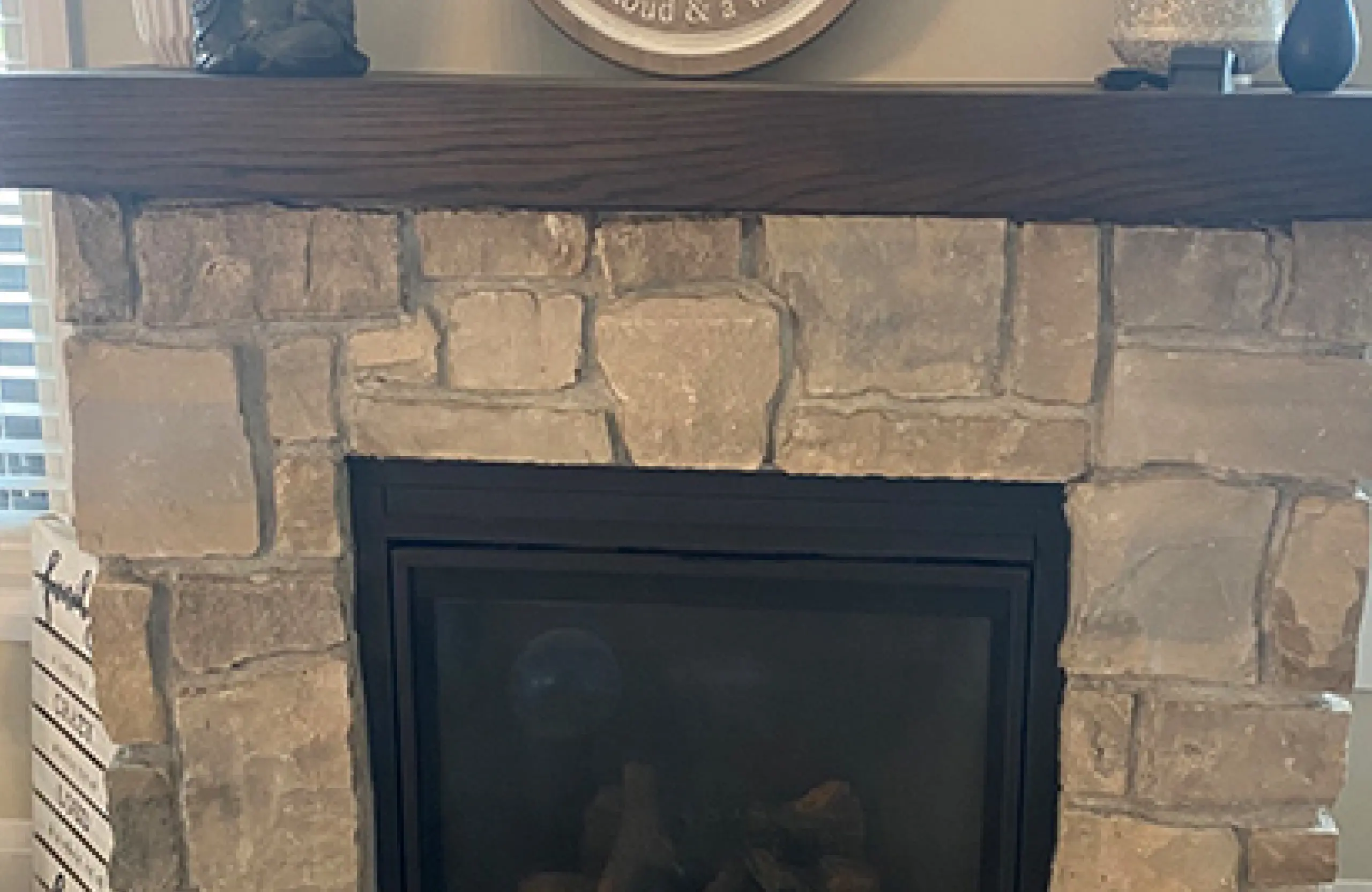 Stone Haven Developments Stone fireplace with a wooden mantel, decorative sign, and small objects displayed on top. Firewood is visible behind the glass front of the fireplace. Ontario