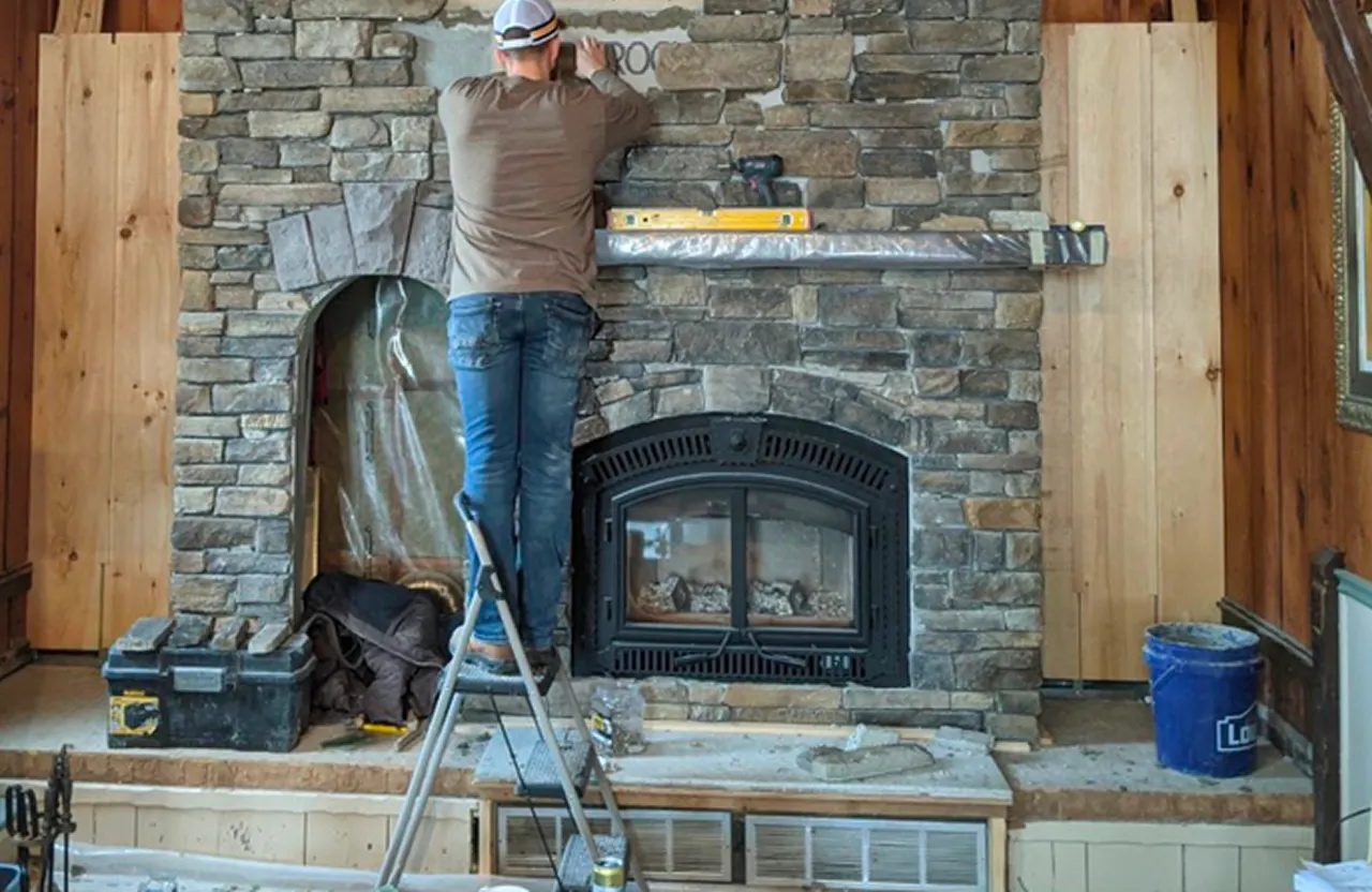 Stone Haven Developments Person standing on a ladder installs or repairs a stone fireplace with tools and construction materials scattered nearby. Ontario