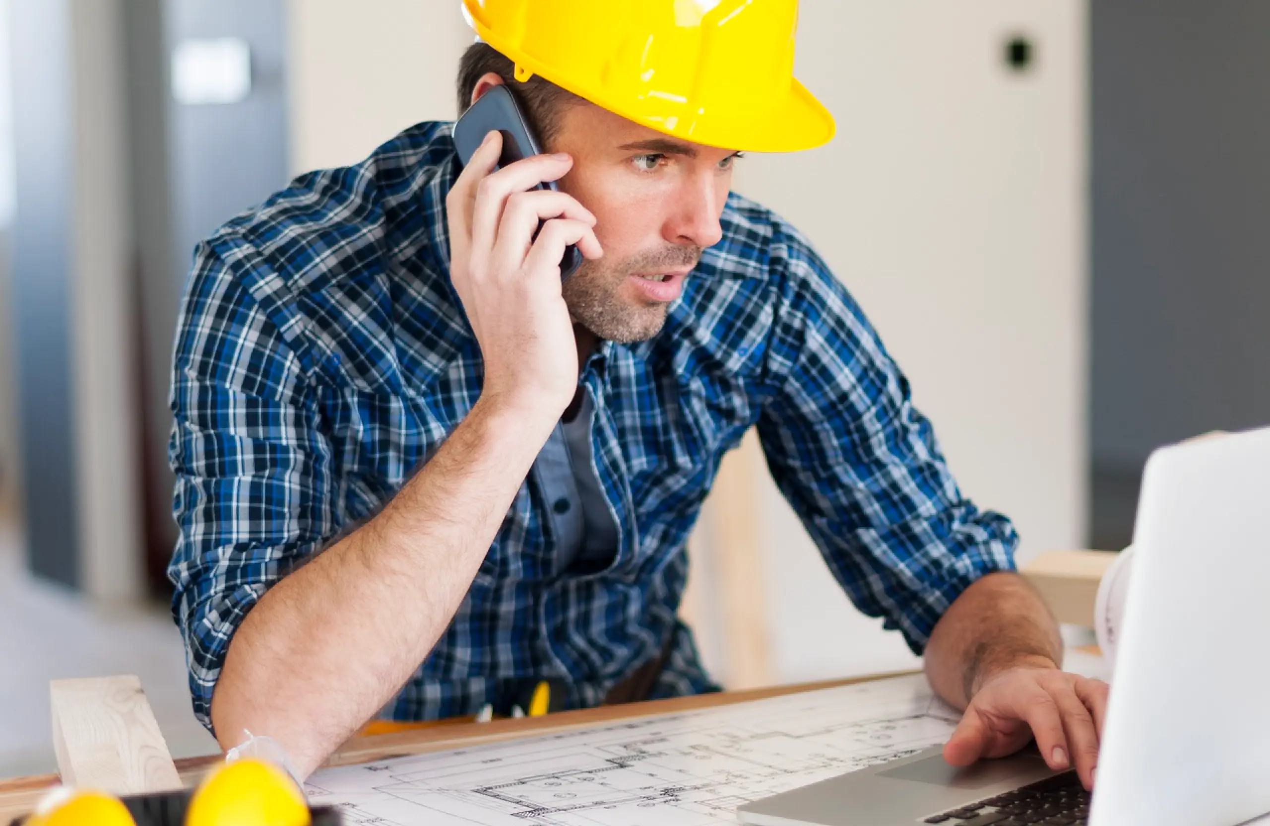 Contractor in a hard hat on the phone while reviewing blueprints and a laptop, part of vetting a contractor in Canada