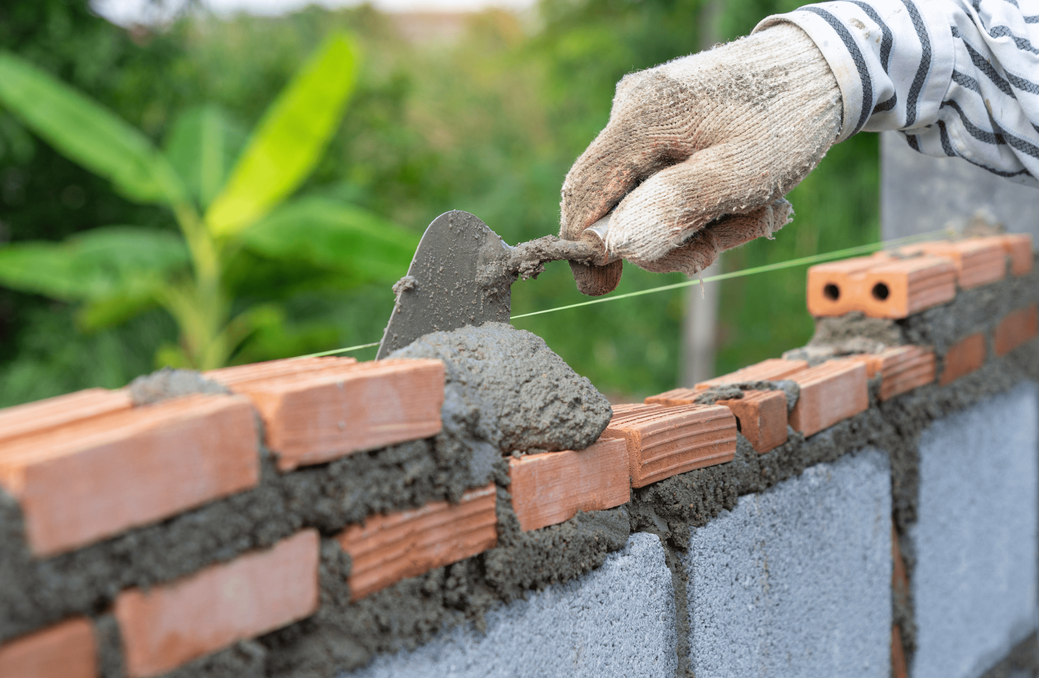 Stone Haven Developments A gloved hand uses a trowel to spread mortar between red bricks while building a wall outdoors, showcasing the craftsmanship of Stone Haven Developments. Ontario